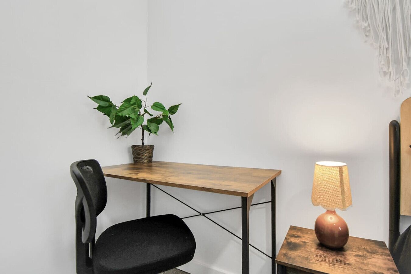 A small workspace with a wooden desk, a black chair, a potted plant, and a table lamp.