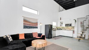Un appartement moderne avec un canapé noir, une table basse ronde en bois et une cuisine dotée de meubles blancs et d’un réfrigérateur noir.