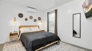 Une chambre à coucher comprenant un lit, des tables de chevet, des éléments de décoration murale, un climatiseur, une fenêtre avec des rideaux et un miroir.