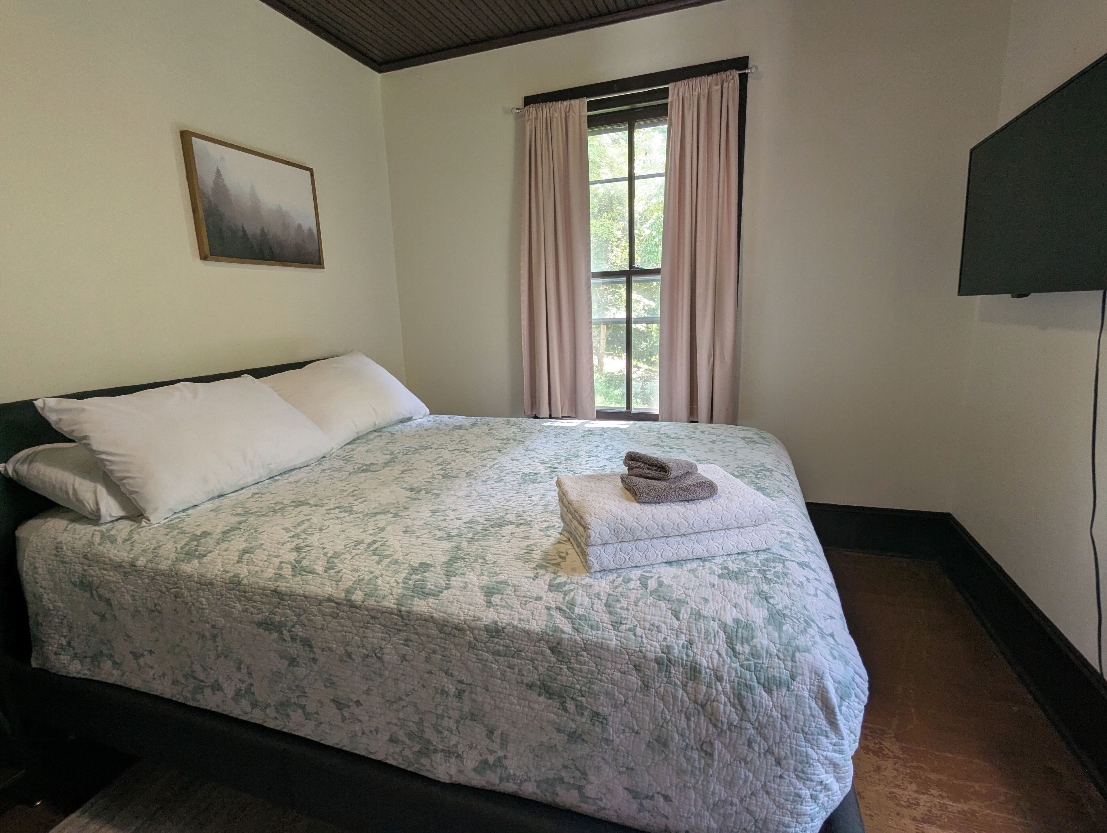 A bedroom with a bed, a window with curtains, a painting on the wall, and a TV mounted on the wall.