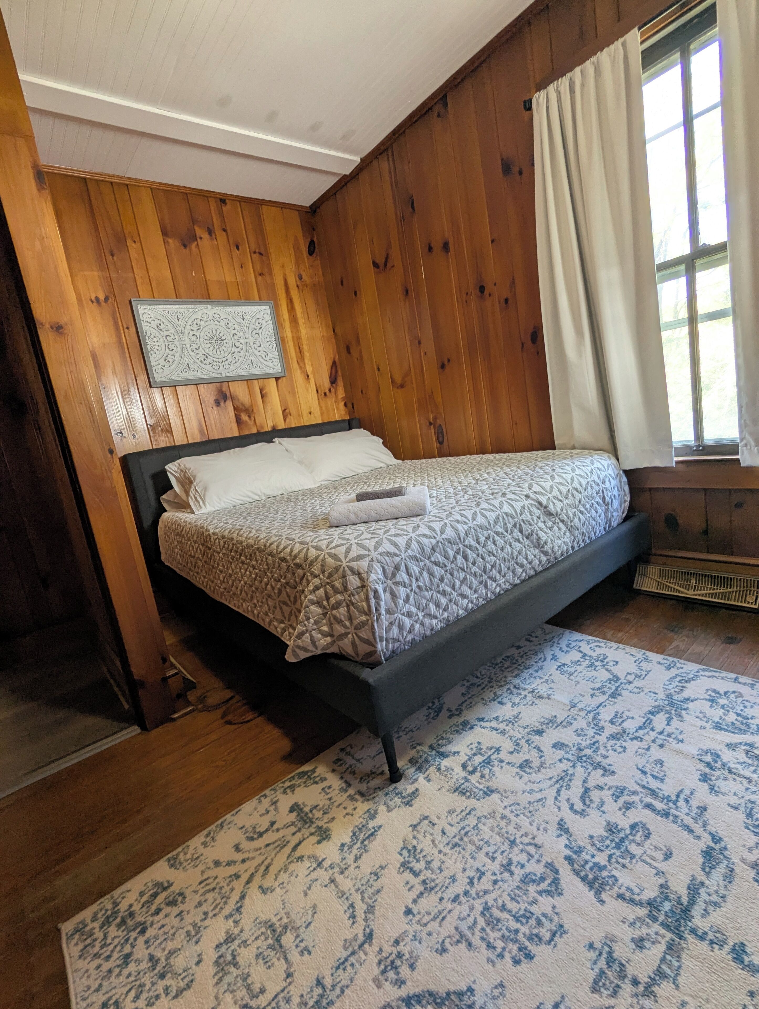 A bedroom with a bed, wooden walls, a window with curtains, and a patterned rug.