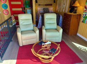 Una habitación acogedora con dos sillones color beige, una mesita redonda con prismáticos, una alfombra roja y una cama con ropa de cama estampada.