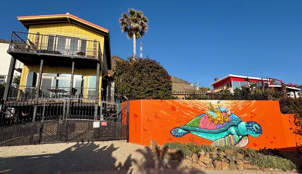 Un edificio con fachada amarilla y un balcón, una palmera y un mural colorido en una pared.