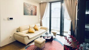 A modern living room with a sofa, ottoman, and coffee table. There is a large window with curtains, a ceiling light, and a wall-mounted artwork. - Sunny - Vinhomes Skylake Residences (Hanoi)