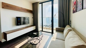 A modern living room with a flat-screen TV, a glass coffee table, a sofa, and a balcony with a city view. - Sunny - Vinhomes Skylake Residences (Hanoi)