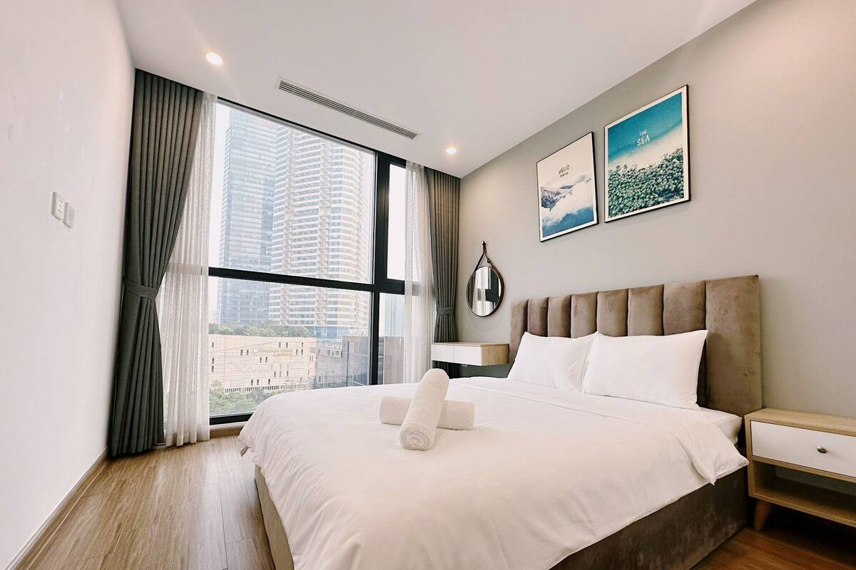 A modern hotel room with a large bed, a bedside table, a mirror, and a view of tall buildings through the window.