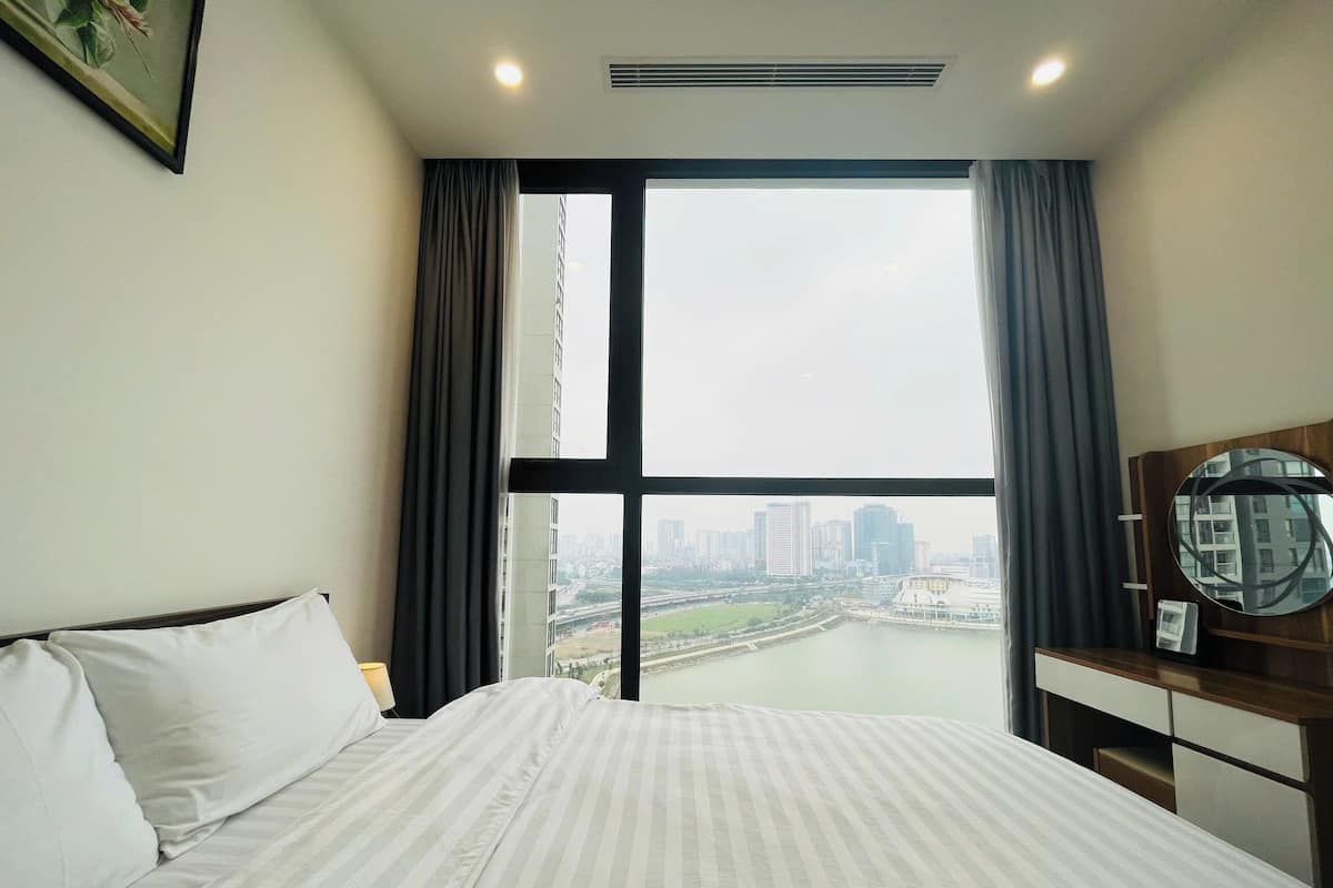 A hotel room with a large bed, a view of a cityscape, and a round mirror on a wooden dresser.