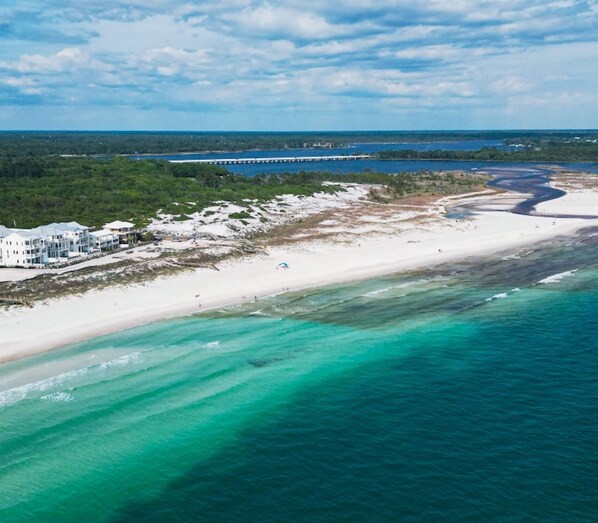 An aerial view of a beach with a white sandy shore, clear turquoise waters, and a few buildings in the background.