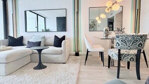 A modern living room with a white sofa, a round coffee table, and a patterned armchair. There is a dining area with a white table and chairs. The room features striped wallpaper and large mirrors.
