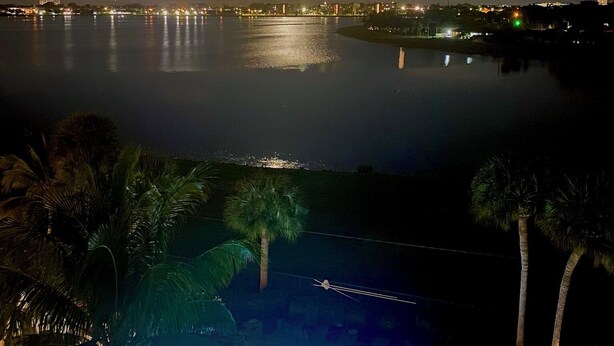 A nighttime view of a pool area with a moonlit sky, a calm body of water reflecting lights, and a distant cityscape.