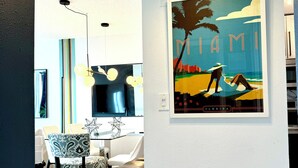 A modern living room with a flat-screen TV, a dining area with patterned chairs, and a wall-mounted painting of a beach scene.