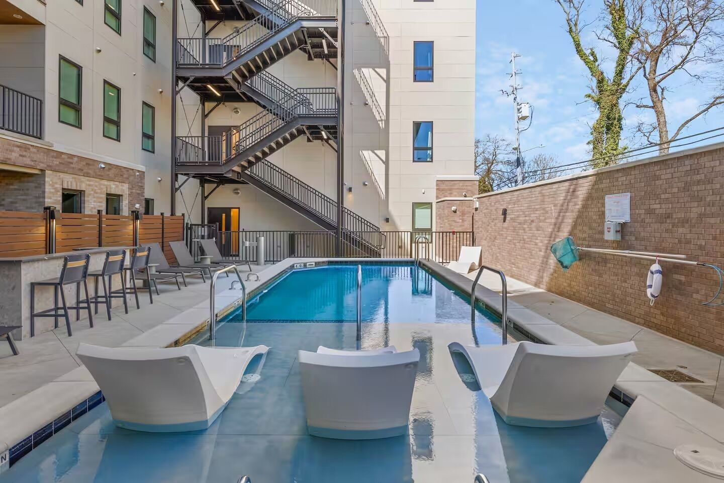 A modern indoor pool with a staircase leading to an upper floor, surrounded by lounge chairs and a brick wall.