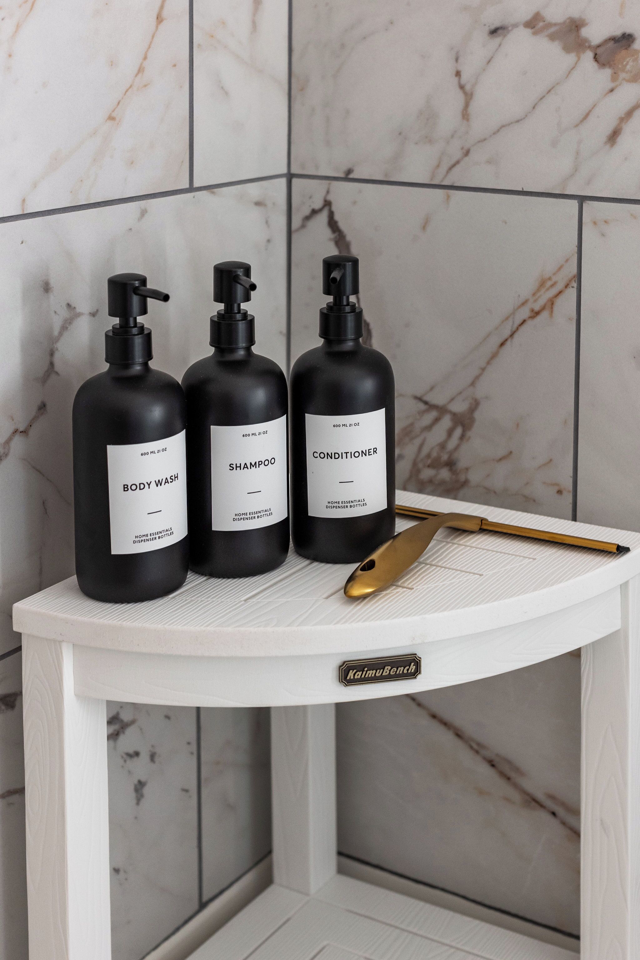 Three black bottles of body wash, shampoo, and conditioner on a white shelf against a marble wall.