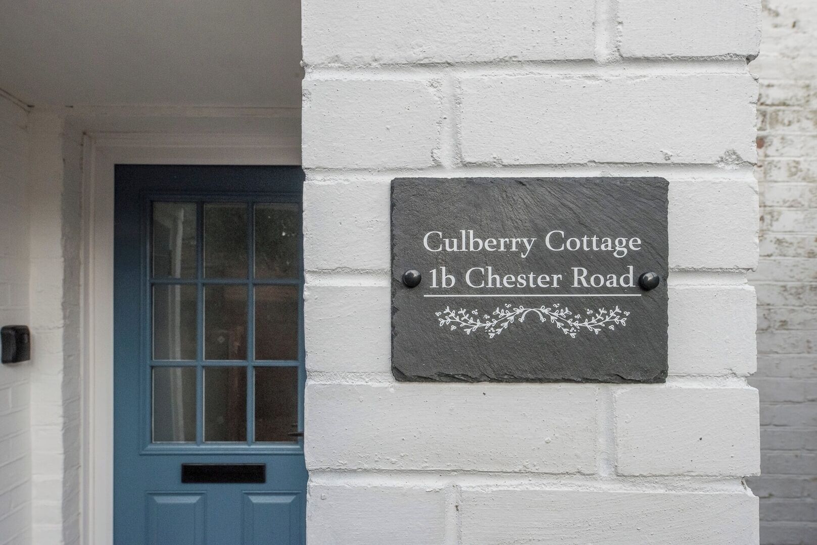 A slate sign with 'Culberry Cottage 1b Chester Road' on a white brick wall.