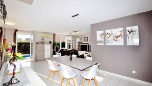 A modern dining area with a long table and white chairs, a kitchen in the background, and artwork on the walls. - Lovely home in Lézignan-Corbières (Lézignan-Corbières)