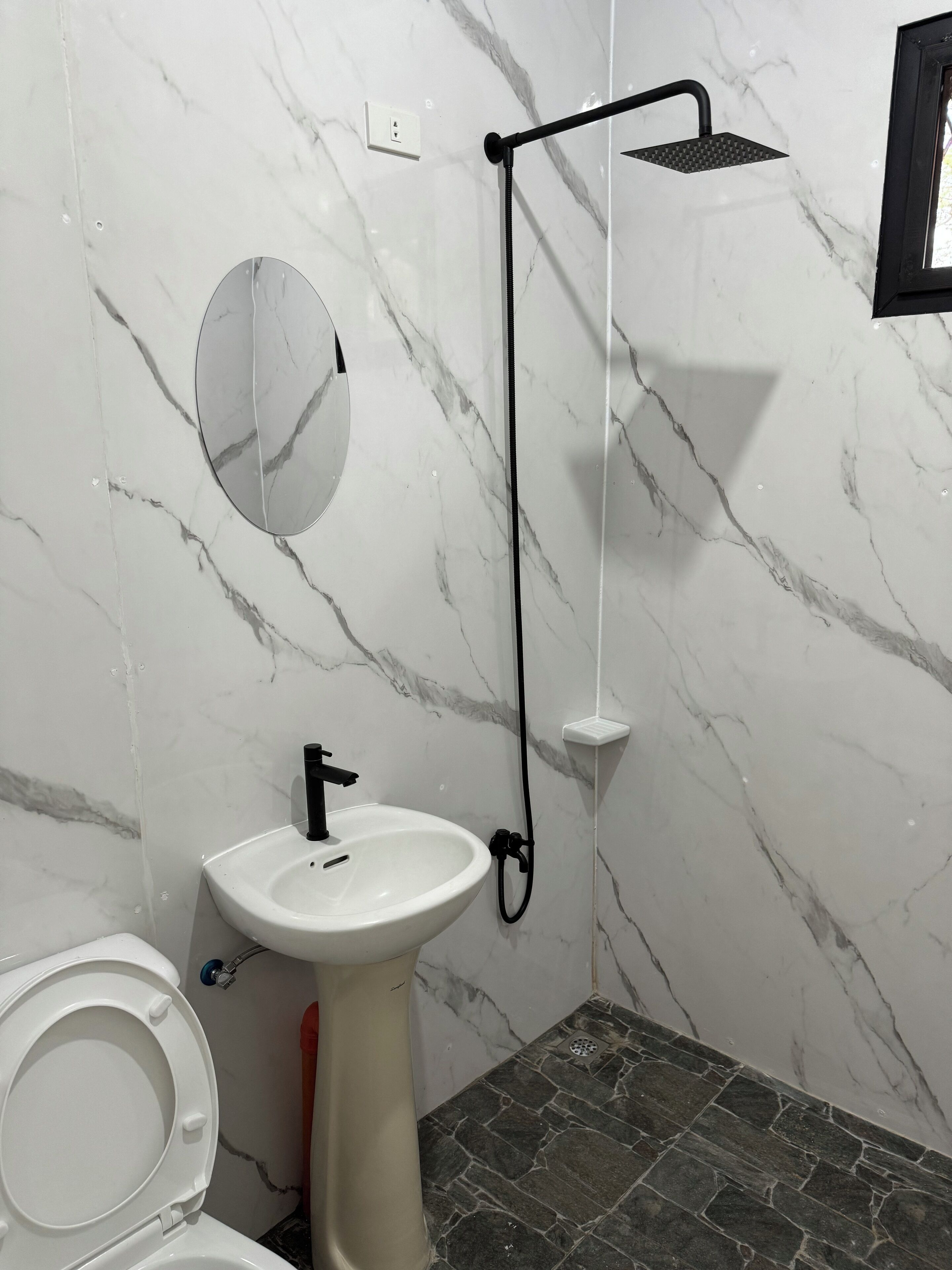 A bathroom with a white marble wall, a pedestal sink, a toilet, and a shower with a black hose.