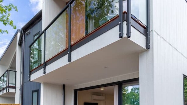 A modern two-story house with a balcony, a wooden deck, and a small outdoor seating area with two white chairs. - The Ultimate 4-Bedroom City Escape (Christchurch)