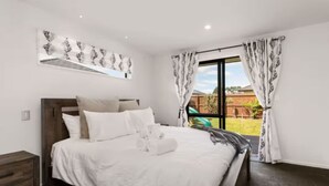 Ein Schlafzimmer mit Bett, Nachttisch, Fenster mit Vorhängen und Blick ins Freie.