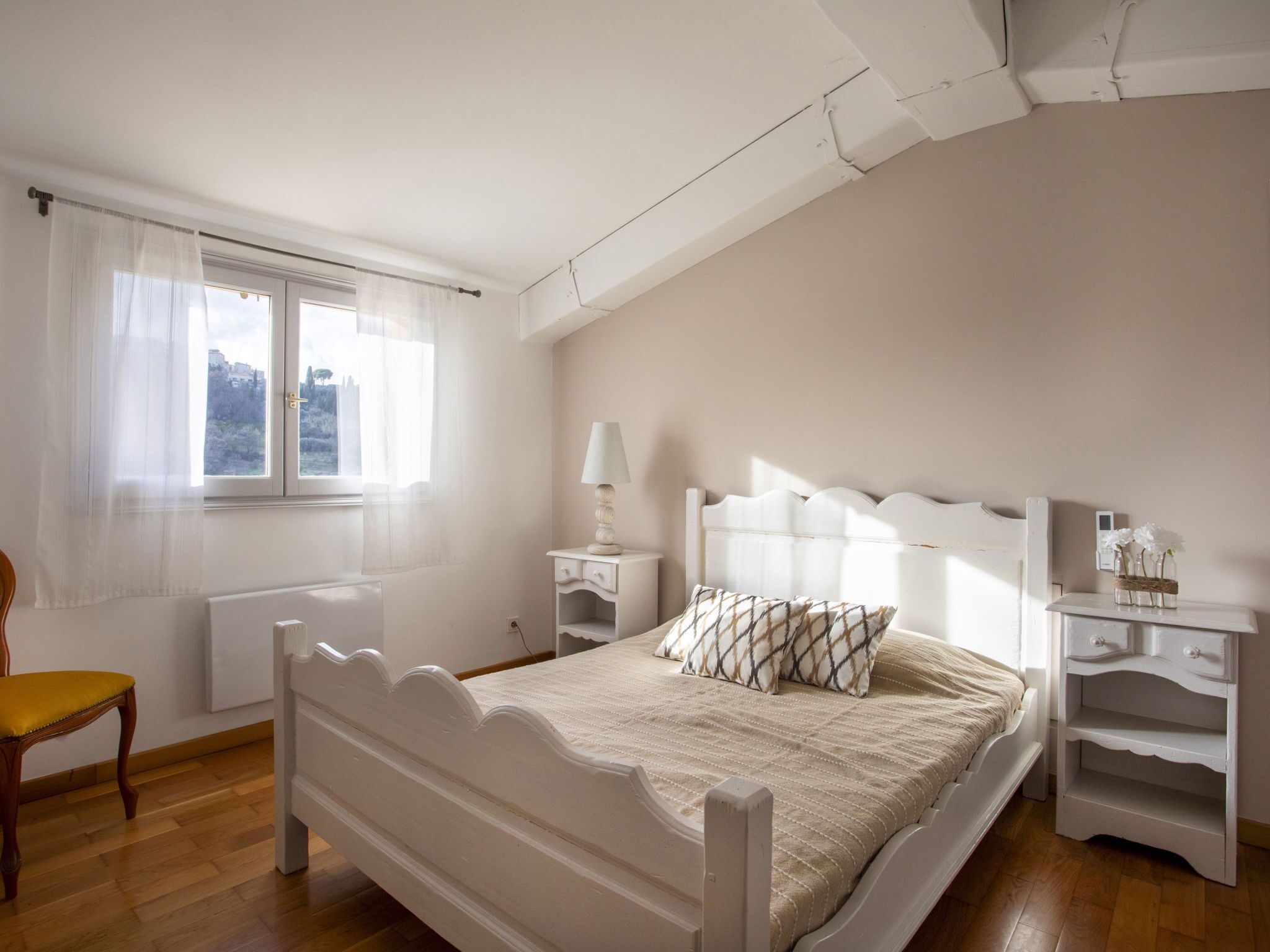 Une chambre à coucher comprenant un lit, une chaise, une table de chevet, une lampe et une fenêtre avec des rideaux.