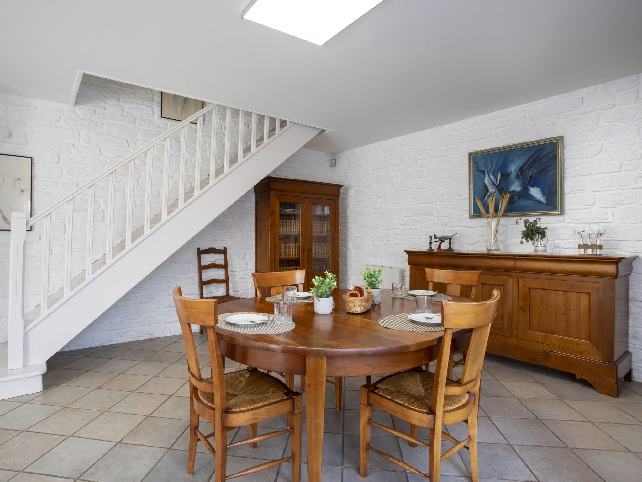 Un espace repas avec une table et des chaises en bois, un escalier, une cheminée et un mur en briques blanches.