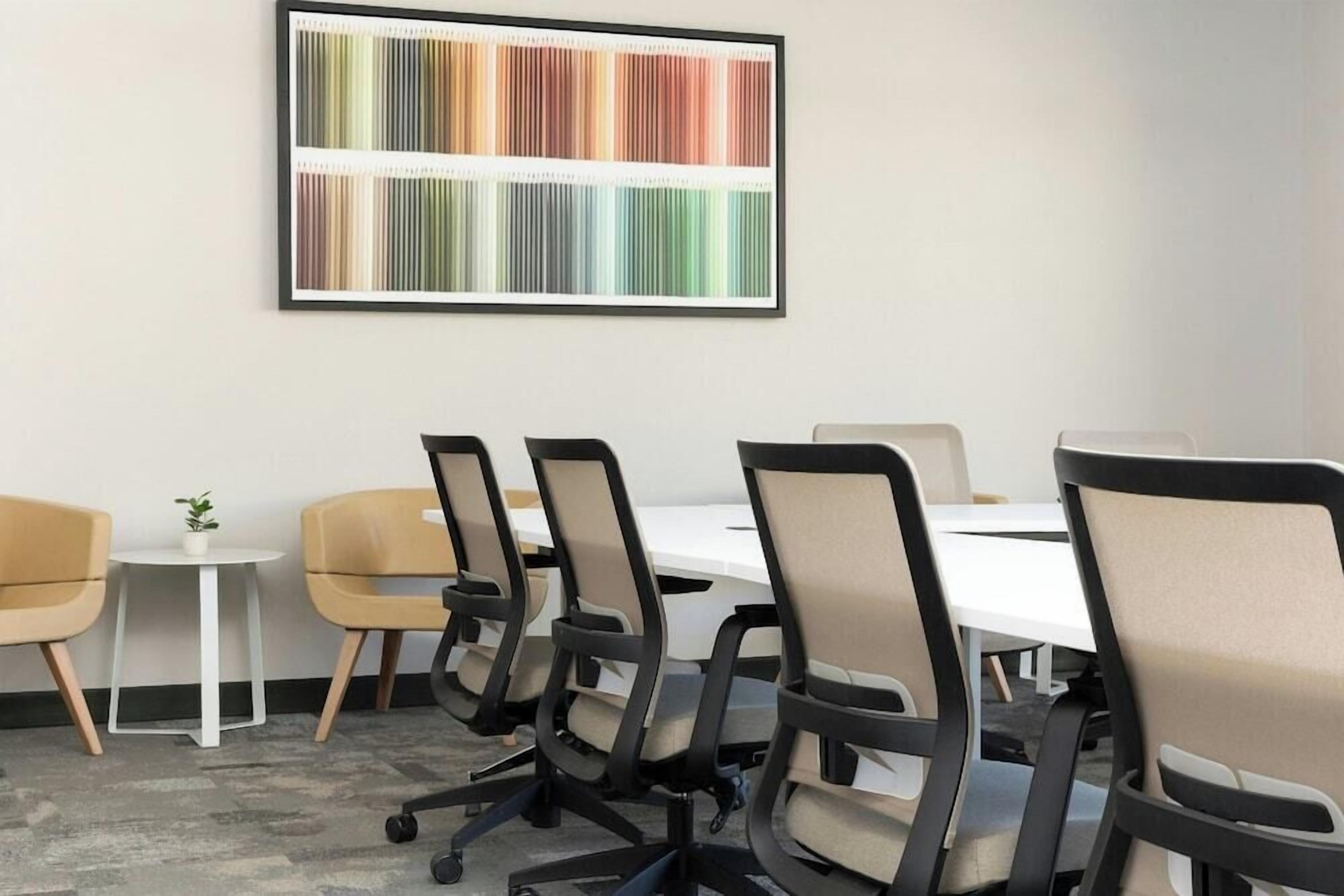 Uma sala de conferências com uma mesa branca, seis cadeiras, uma pequena mesa branca com uma planta e um quadro abstrato colorido na parede.