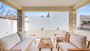 A covered outdoor seating area with two sofas and a wooden table, a stone wall, and a view of a building in the background.