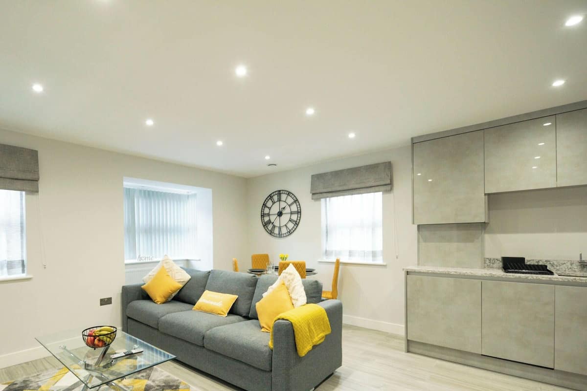 A modern living room with a grey sofa, yellow cushions, a glass coffee table, and a kitchen area with grey cabinets.