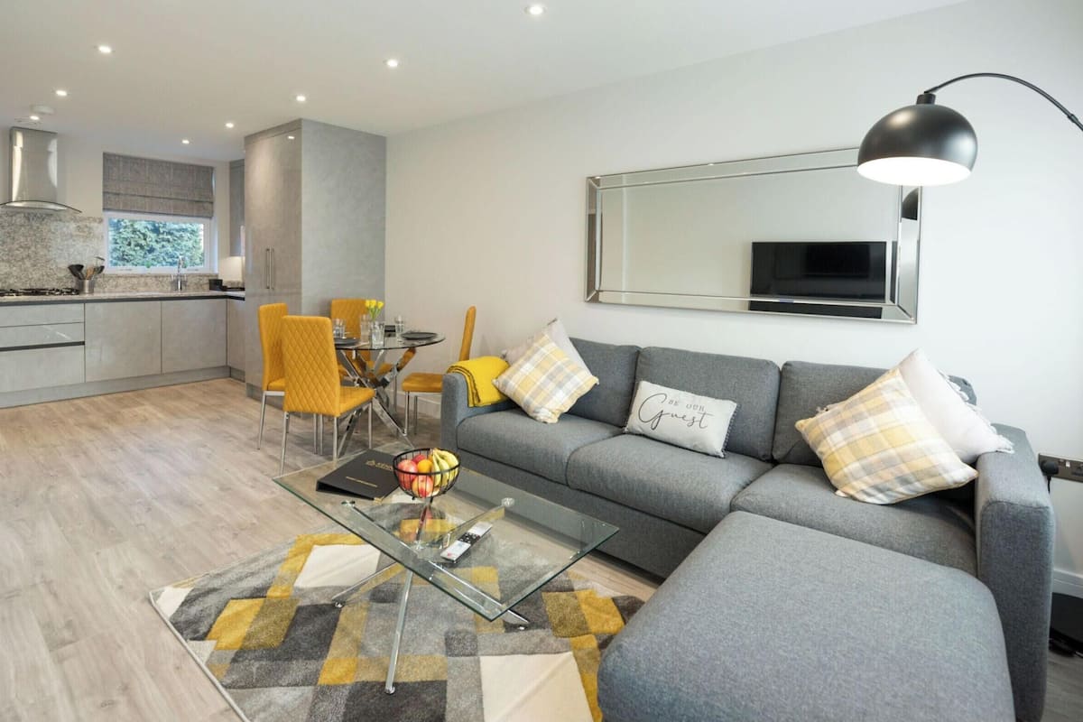 A modern living room with a grey sofa, yellow chairs, a glass coffee table, and a large mirror on the wall.
