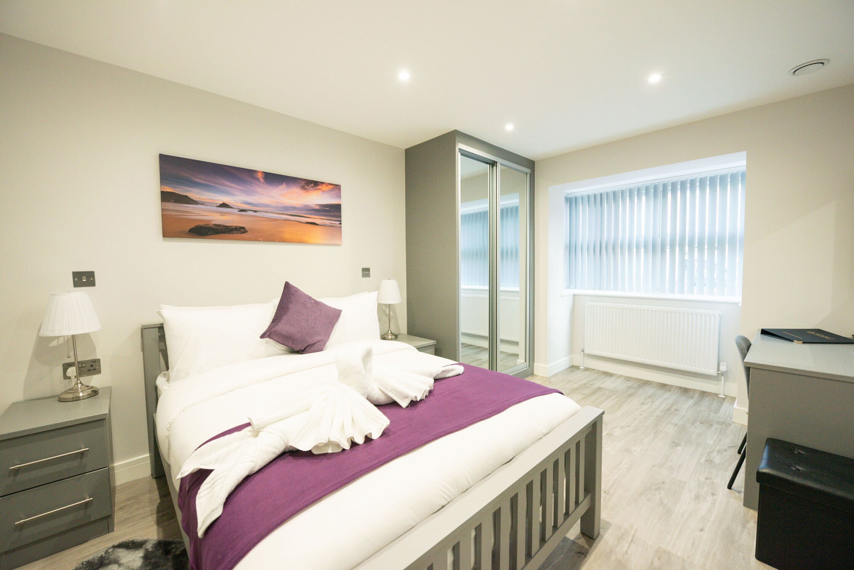 A modern bedroom with a bed, bedside tables, a window with blinds, a radiator, and a painting on the wall.