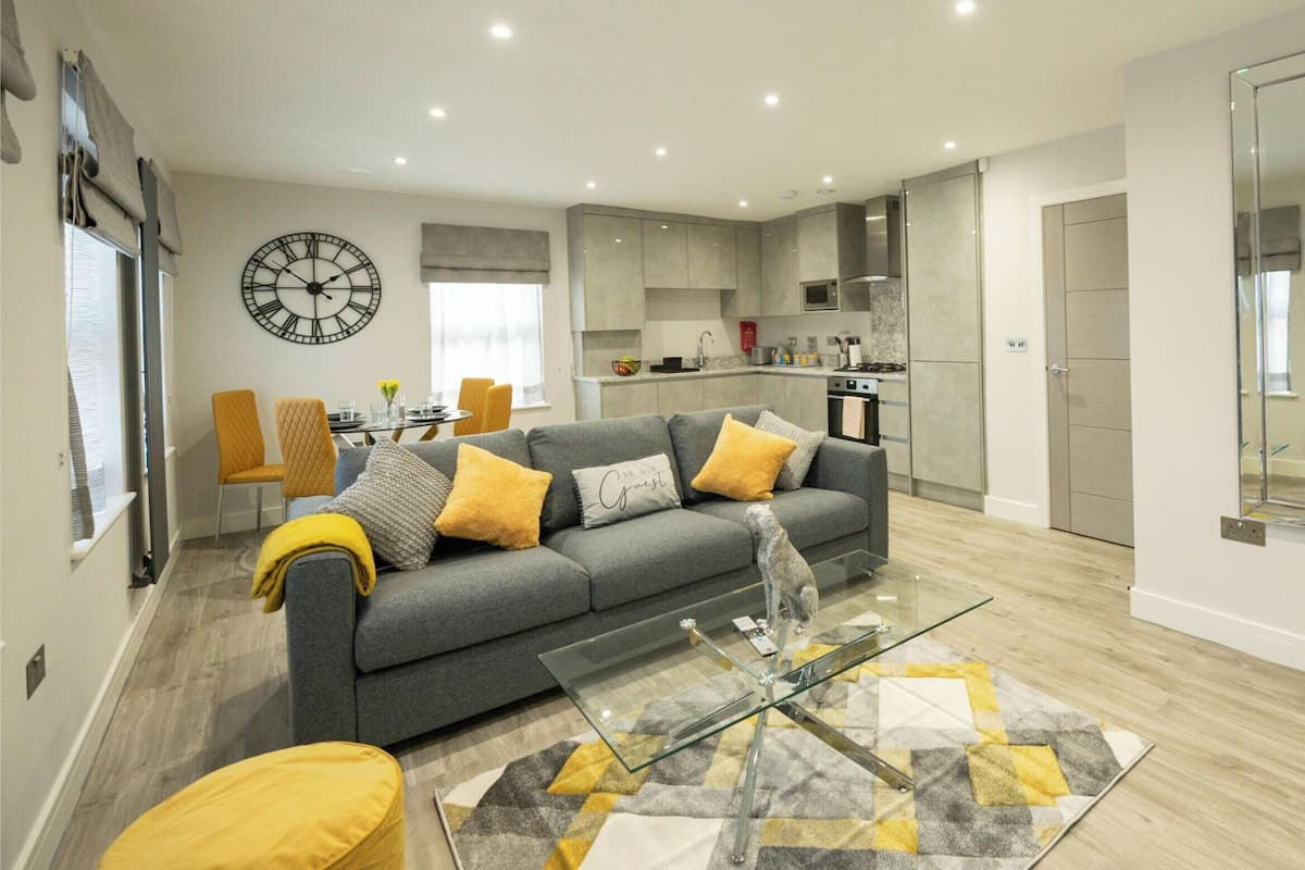 A modern living room with a grey sofa, yellow cushions, a glass coffee table, and a patterned rug.