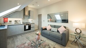 A modern kitchen with a grey countertop, stainless steel appliances, and a built-in oven. There is a grey sofa with pink cushions in the living area, and a glass coffee table. - Chester House (Reading)