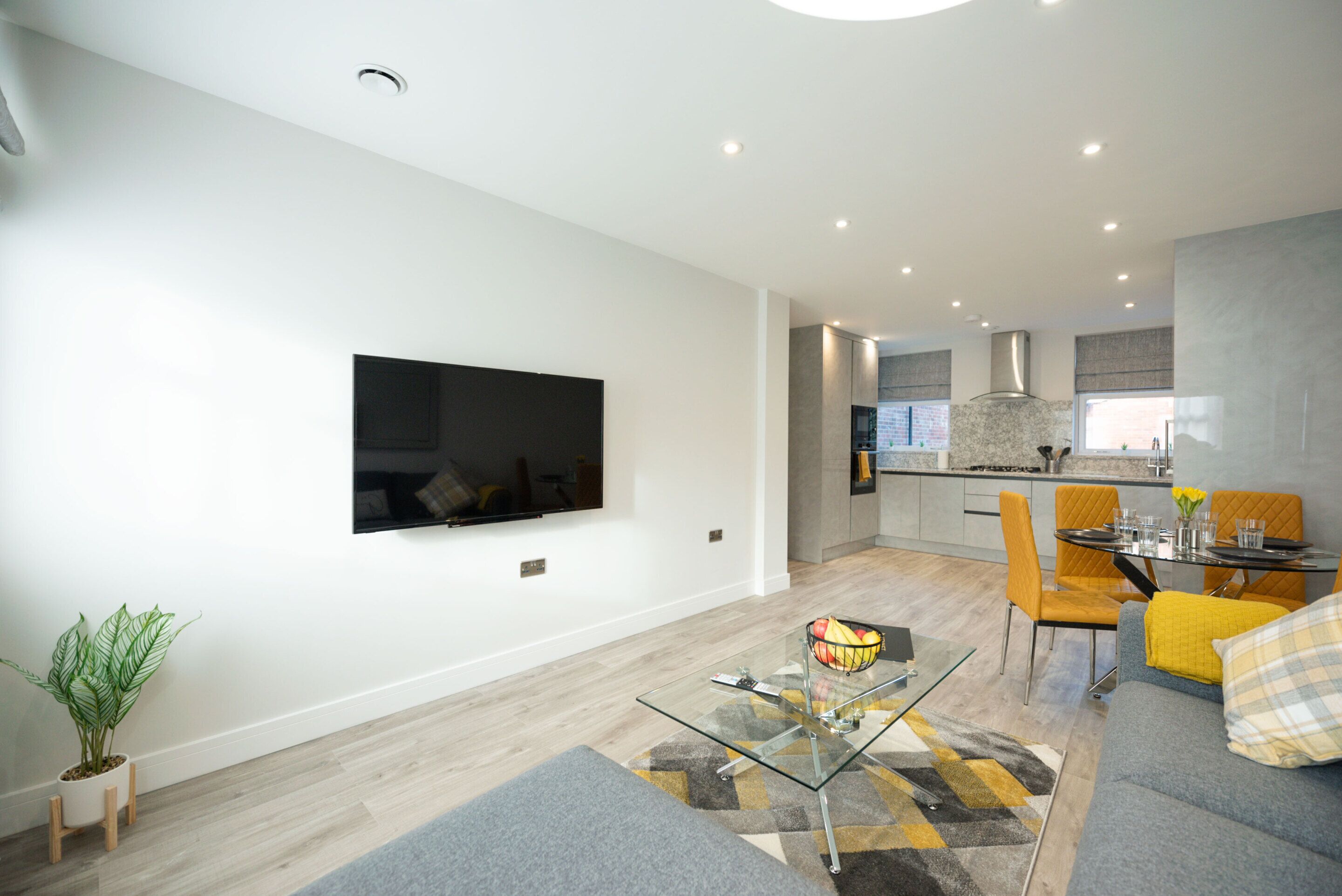 A modern living room with a flat-screen TV, a glass coffee table, a sofa, and a dining area with a table and chairs.