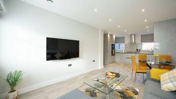 A modern living room with a flat-screen TV, a glass coffee table, a sofa, and a dining area with a table and chairs.