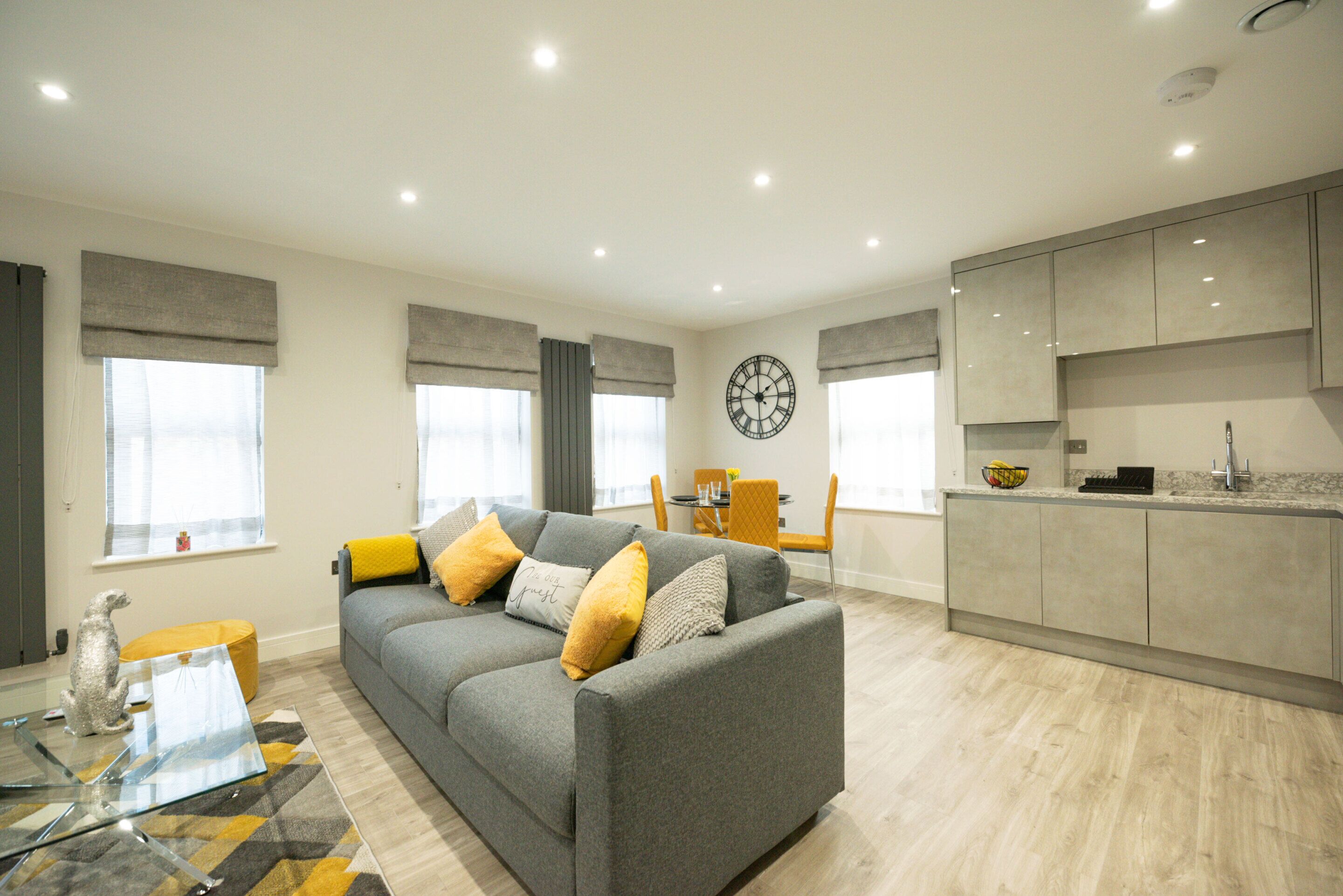 Una sala de estar moderna con un sofá gris, cojines amarillos, una mesa de centro de vidrio y un comedor con una silla amarilla.