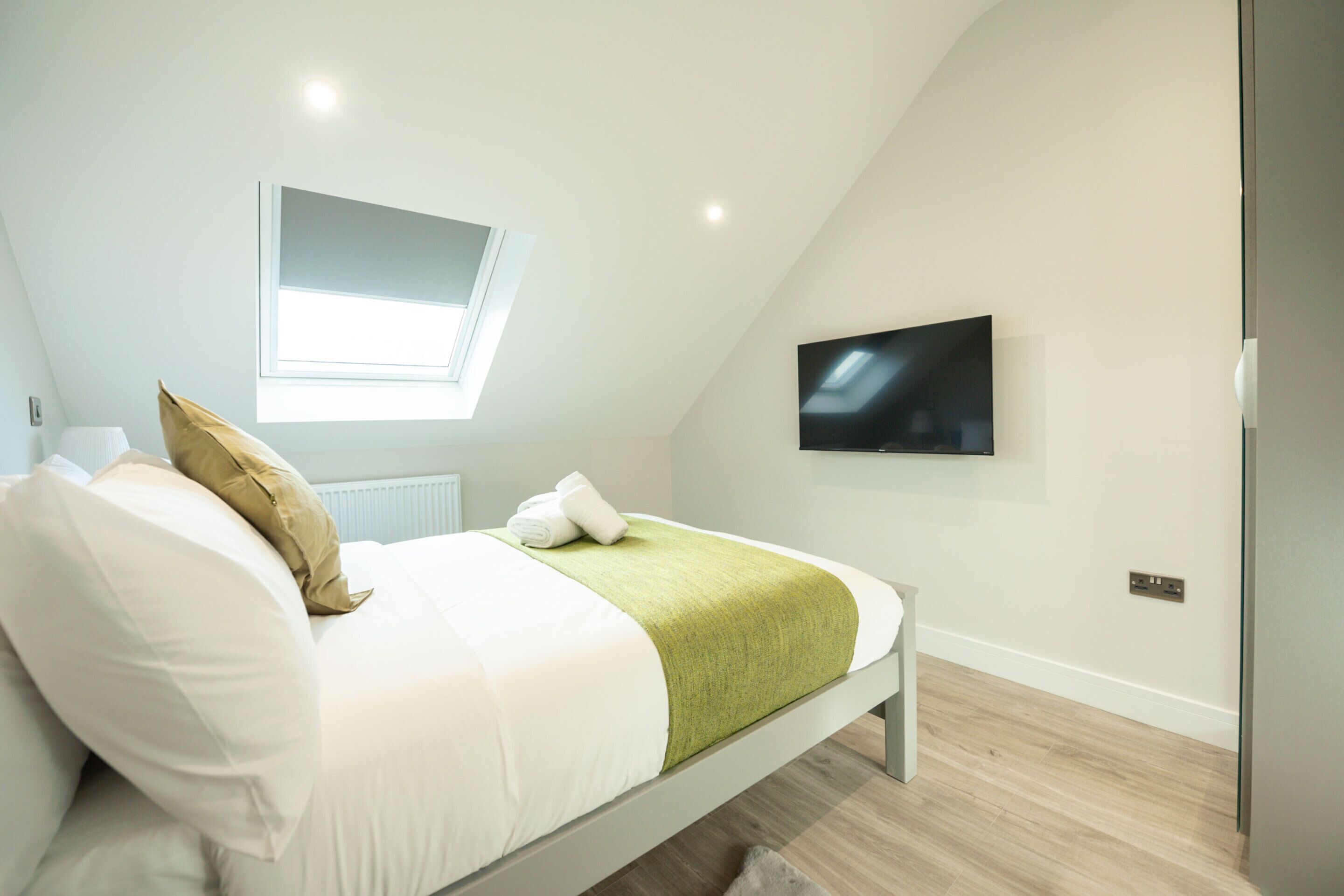 Ein modernes Schlafzimmer mit Bett, Kissen, einer grünen Decke, einem Fenster und einem Fernseher an der Wand.