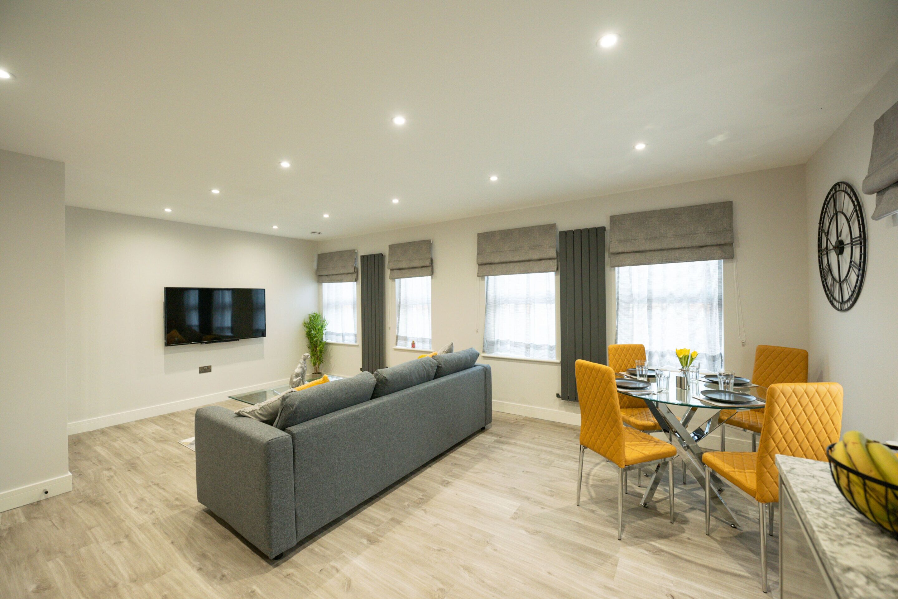 Una sala de estar moderna con un sofá gris, sillas amarillas, una mesa de comedor y un televisor de pantalla plana montado en la pared.
