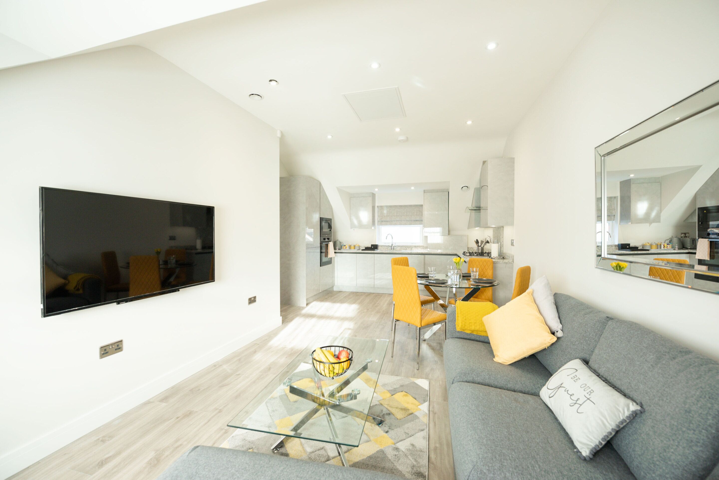 A modern living room with a flat-screen TV, a glass coffee table, a grey sofa, and a dining area with yellow chairs.