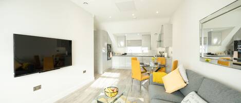 A modern living room with a flat-screen TV, a glass coffee table, a grey sofa, and a dining area with yellow chairs.
