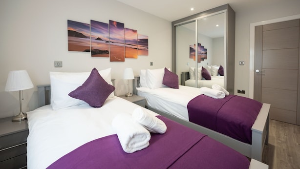 A hotel room with two beds, purple bedding, white pillows, a large mirror, and a sunset painting on the wall. - Chester House (Reading)