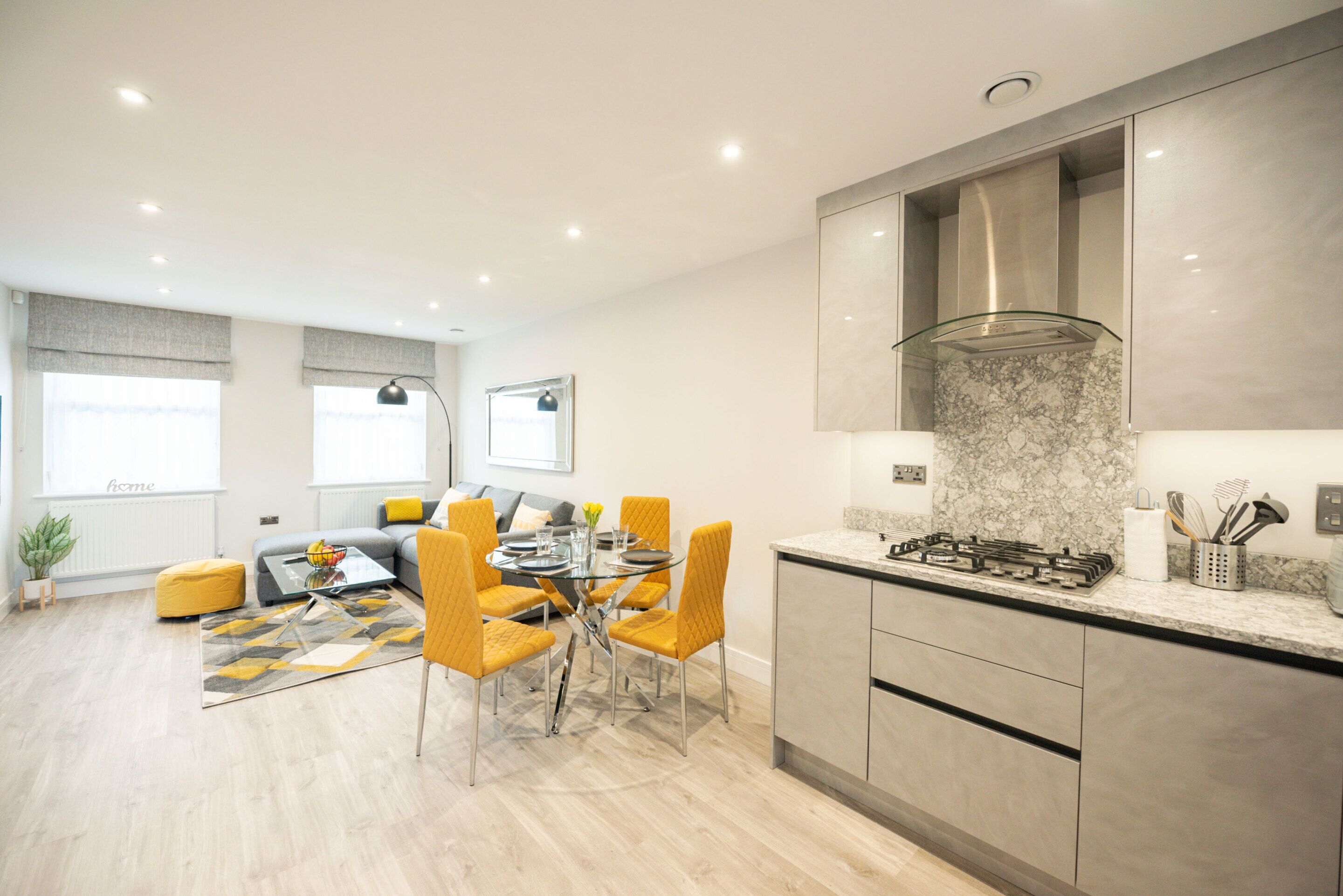 A modern kitchen with a gas stove, marble countertop, and stainless steel cabinets. There is a dining area with a glass table and yellow chairs.