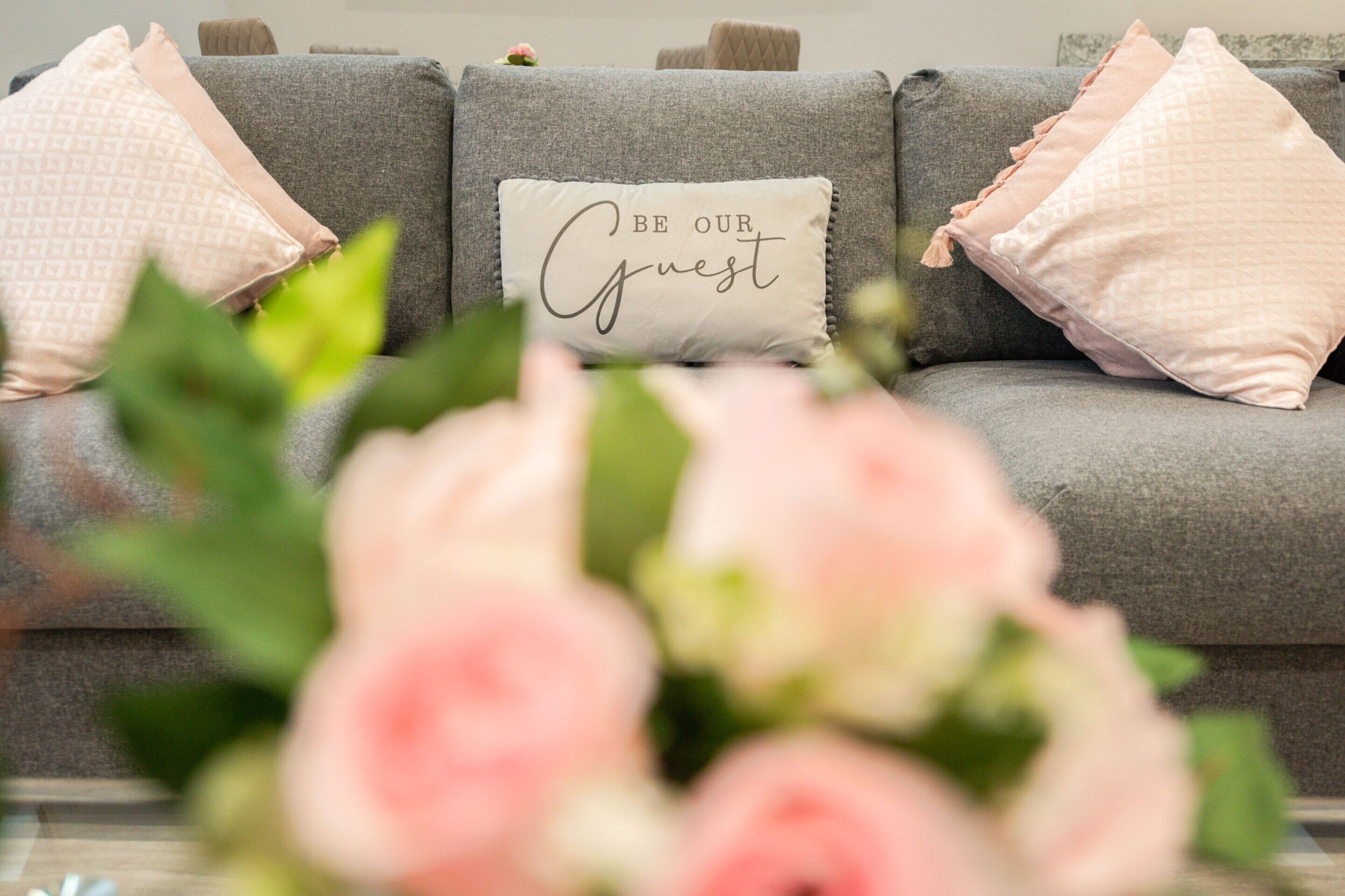 A grey sofa with three pillows, one with the text "BE OUR Guest".