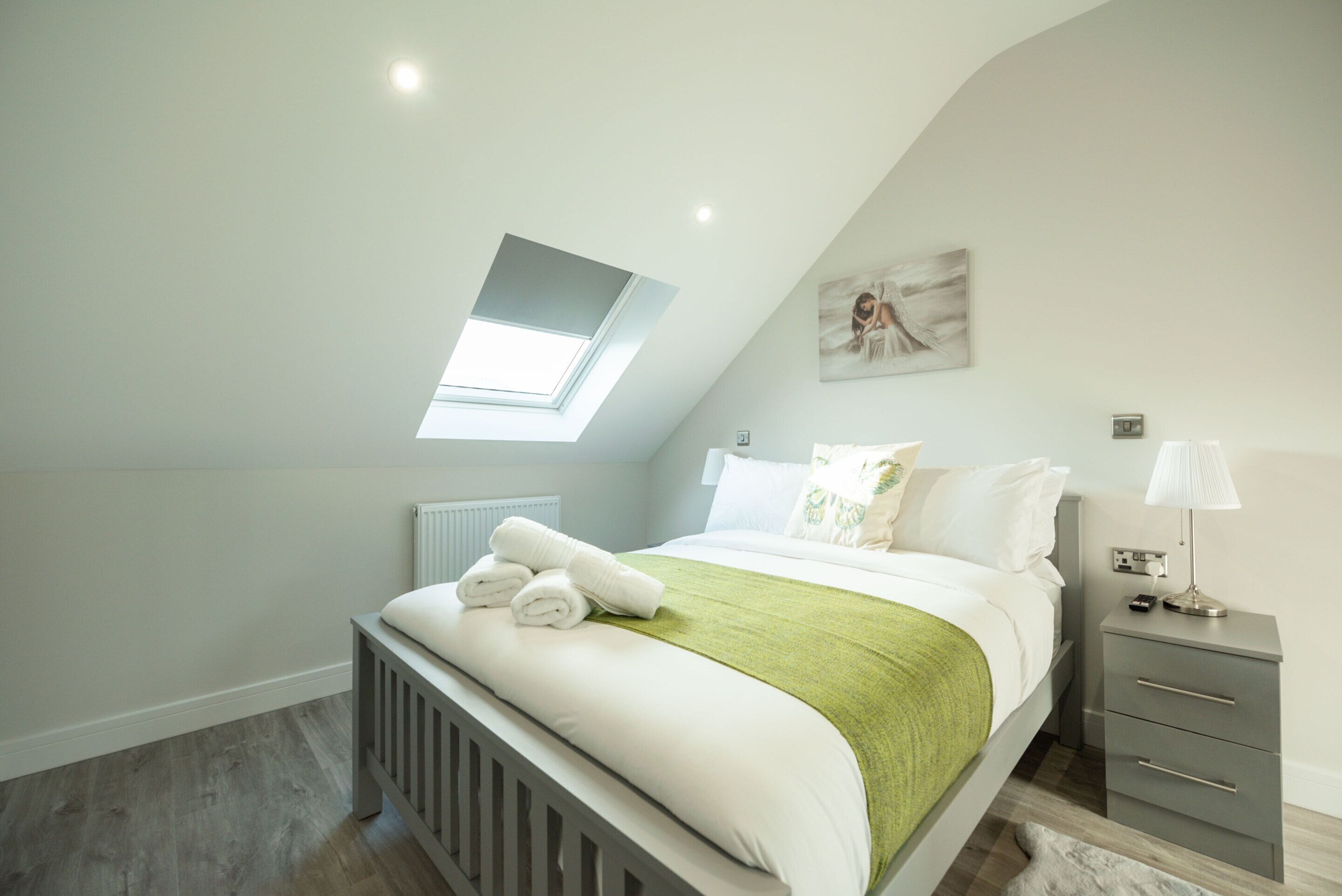 Een slaapkamer met een schuine kap, een dakraam, een bed met wit beddengoed en een groene plaid, een nachtkastje met een lamp, en een radiator.