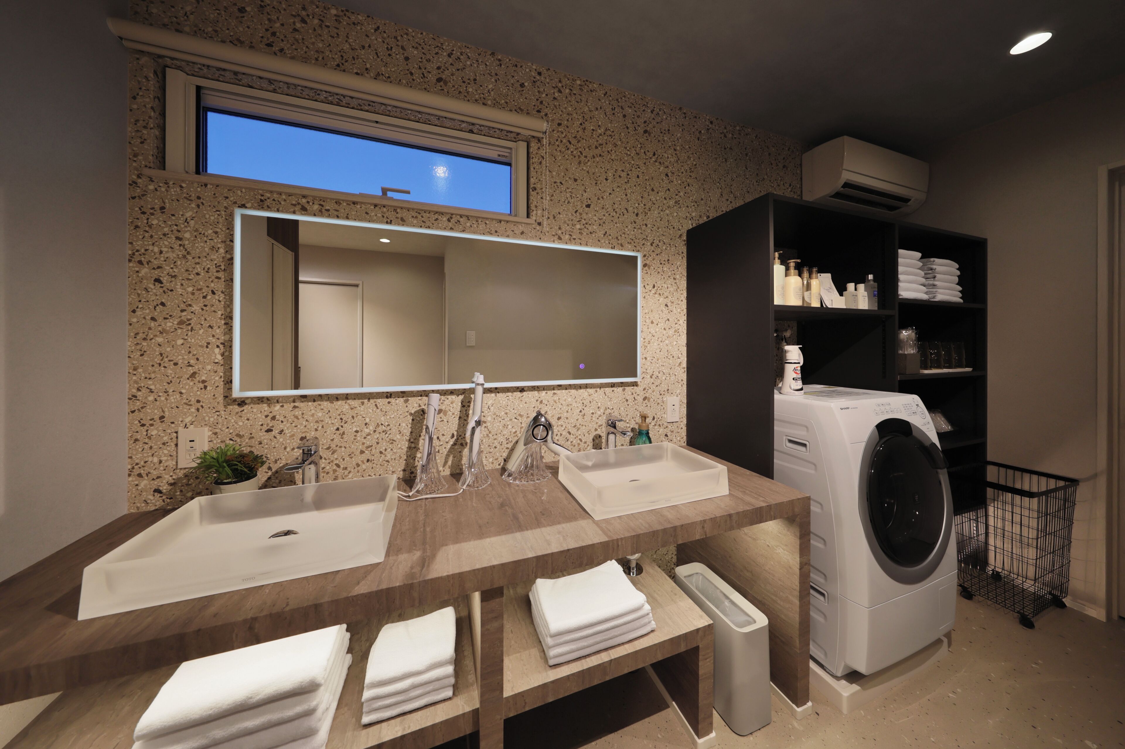 Een moderne badkamer met een grote spiegel, twee wastafels, een ingebouwde wasmachine en een plank met toiletartikelen.