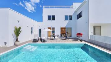 Een modern buitenzwembad met een helderblauwe lucht, een wit gebouw met een balkon en een barbecue.