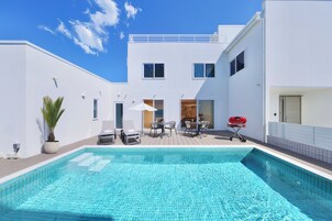 Un área de piscina exterior moderna con cielo azul despejado, un edificio blanco con balcón y una parrilla para barbacoa.