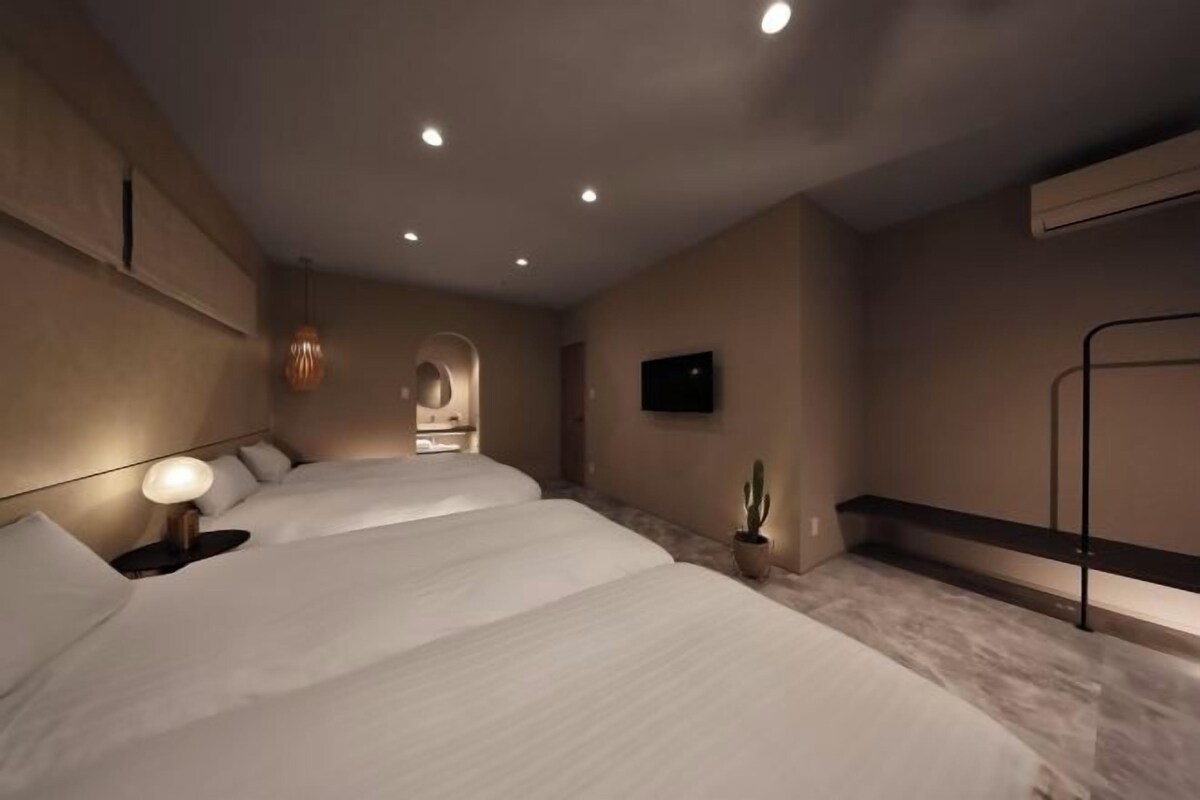 A modern hotel room with three beds, a flat-screen TV, a small cactus plant, and a bench along the wall.
