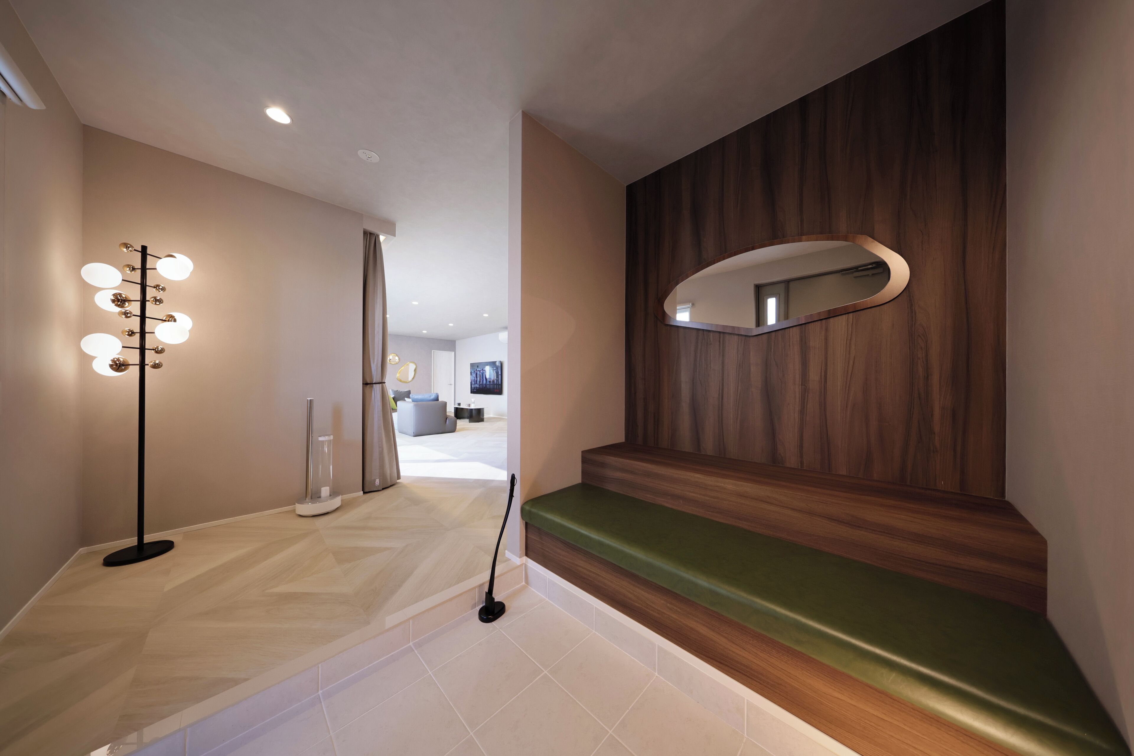 A modern hotel lobby with a light-colored floor, a green cushioned bench, a wall-mounted mirror, and a floor lamp with multiple lights.