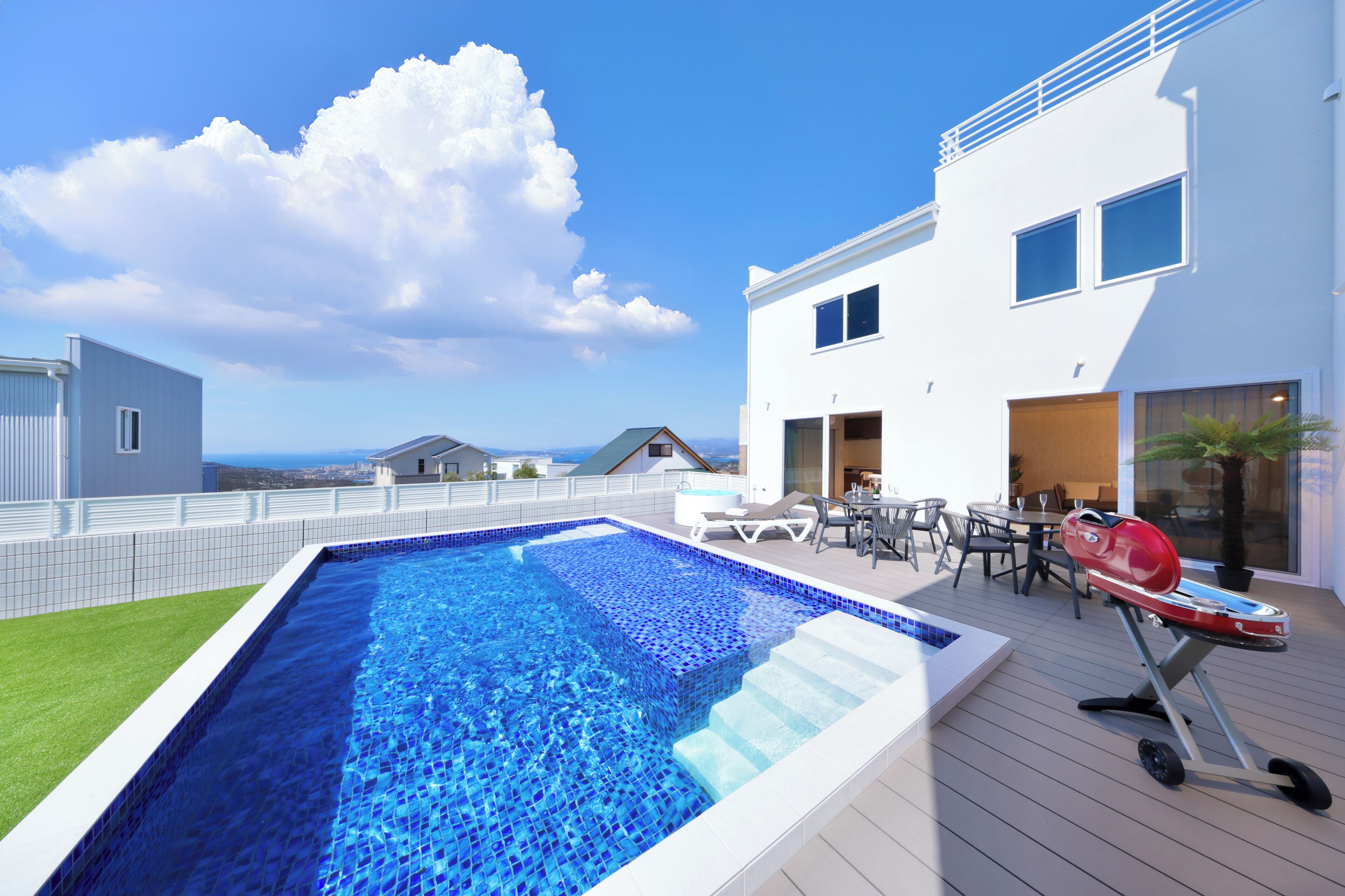 A modern outdoor pool area with a clear blue sky, a wooden deck, and a barbecue grill.