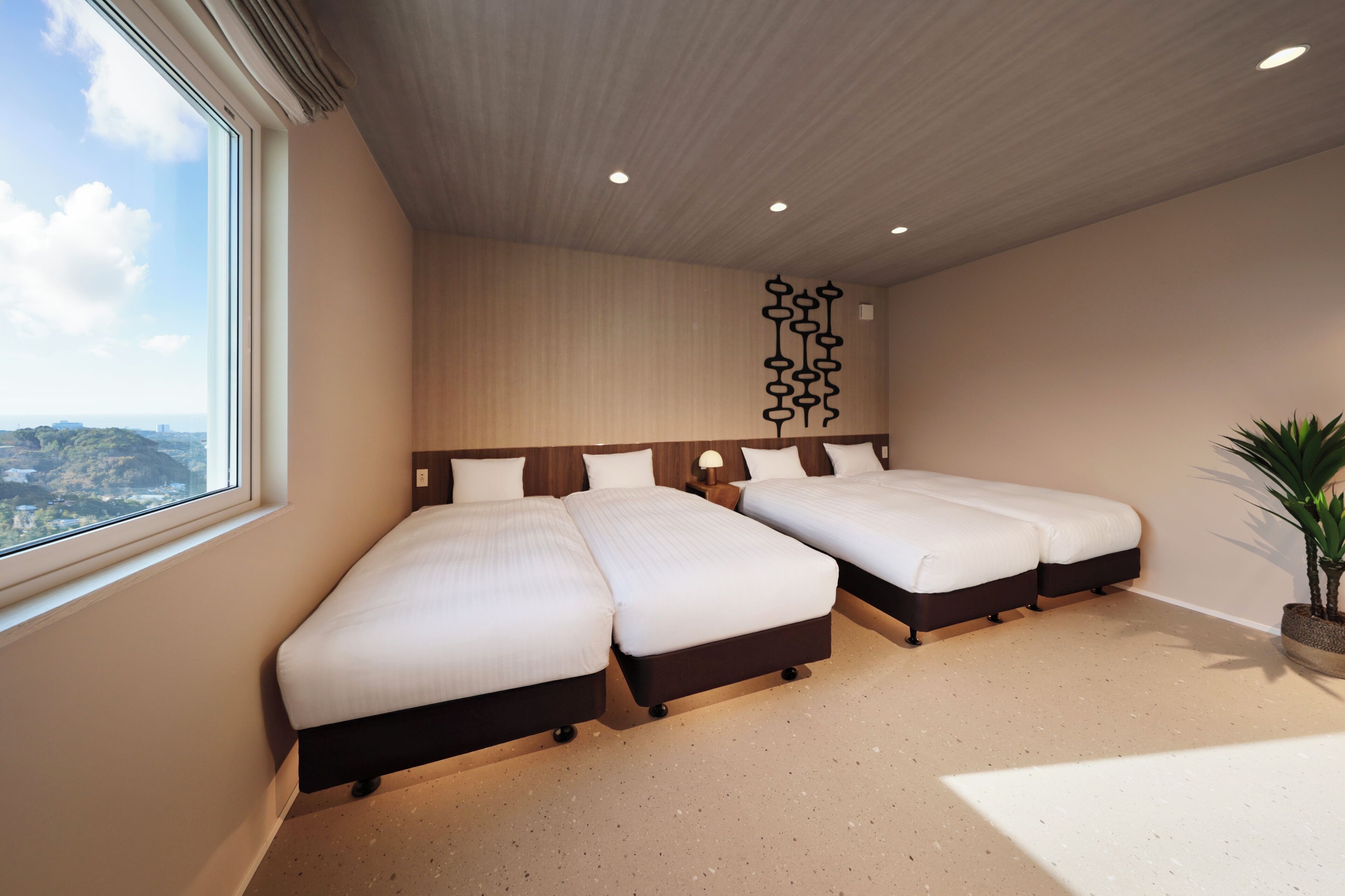 A hotel room with three beds arranged in a row, a large window with a view, a ceiling with recessed lighting, and a decorative wall piece.