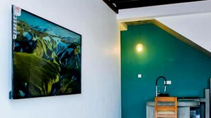 A room with a large abstract painting on the wall, a ceiling fan, a kitchen area with a green wall, and a concrete floor. - Surfside Serenity Ahangama (Ahangama)